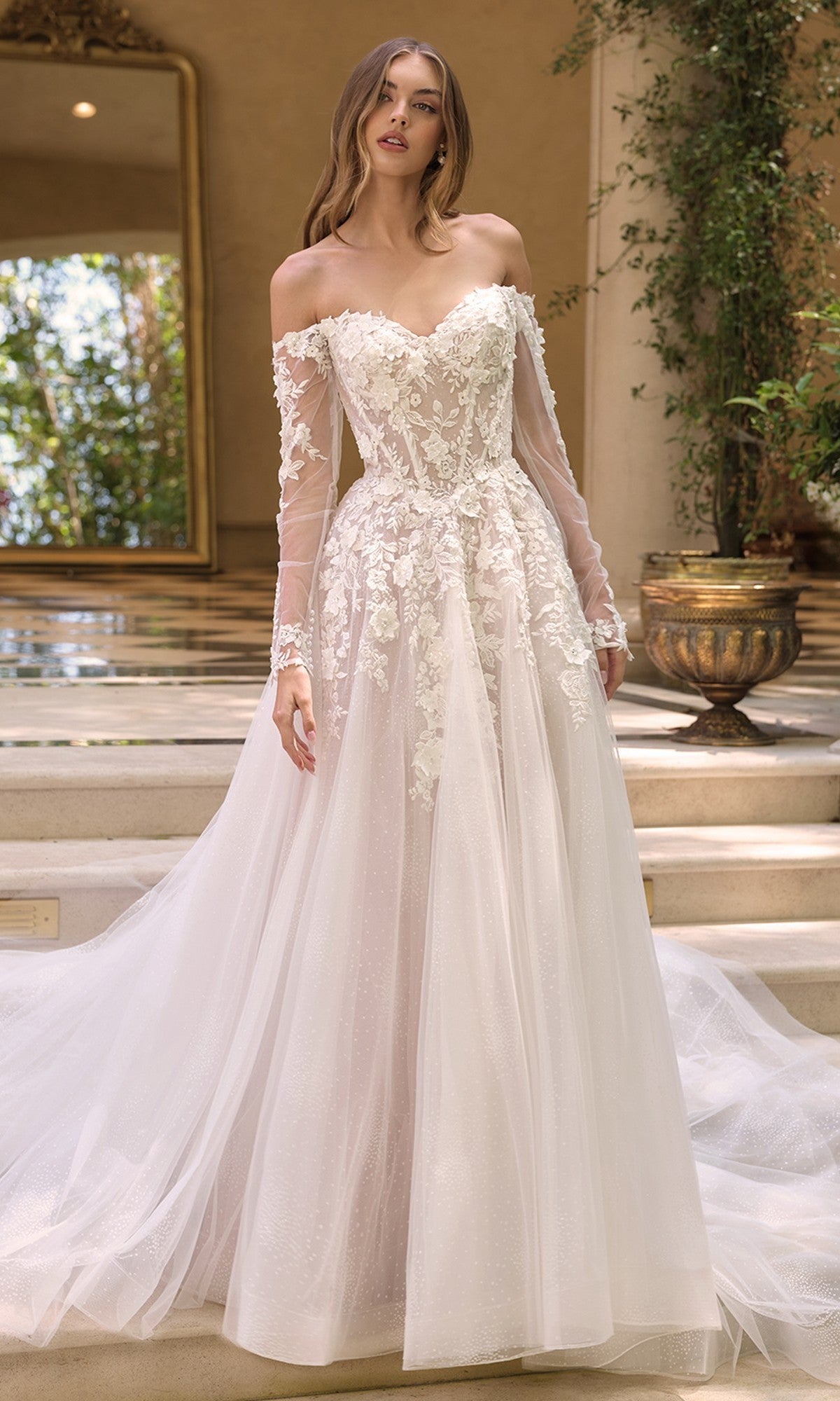 A woman in the Andrea and Leo WL067 white lace, off-the-shoulder wedding dress with long sleeves and a corset bodice stands indoors by steps decorated with greenery, with a large mirror behind her.